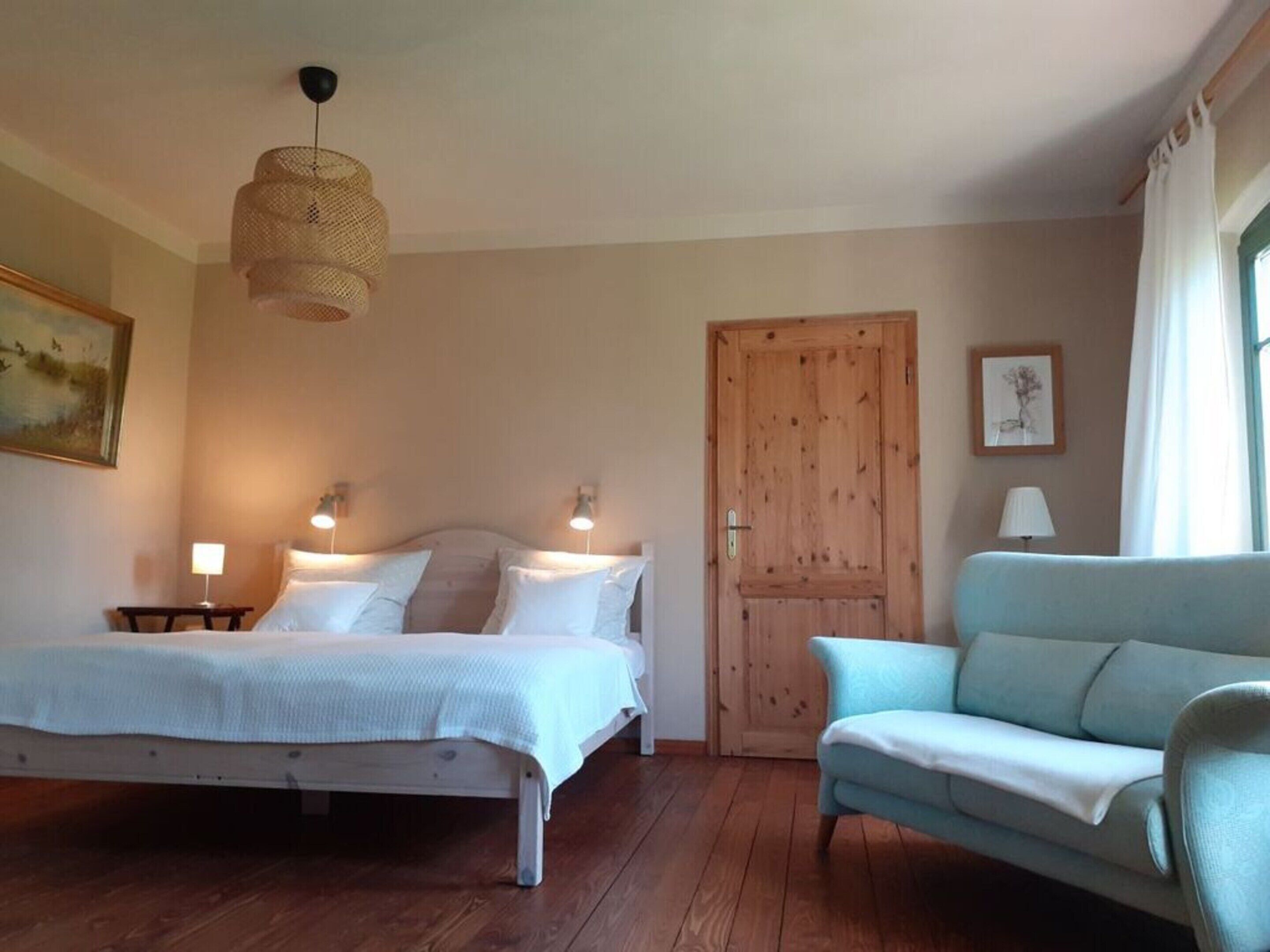 A bedroom with a bed, bedside tables, a hanging light fixture, a wooden door, a blue sofa, and a painting on the wall.