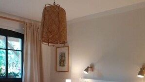 Un grand lit avec du linge de lit blanc, un luminaire suspendu en fibres tressées et une fenêtre avec des rideaux.