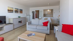 Uma sala de estar moderna com um sofá cinza, almofadas vermelhas, uma mesa de centro com revistas, uma TV e uma área de cozinha ao fundo.