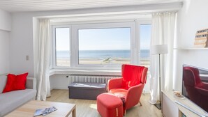 Quarto moderno com uma grande janela com vista para uma praia, uma poltrona vermelha, um sofá cinzento e uma televisão sobre uma mesa de madeira.