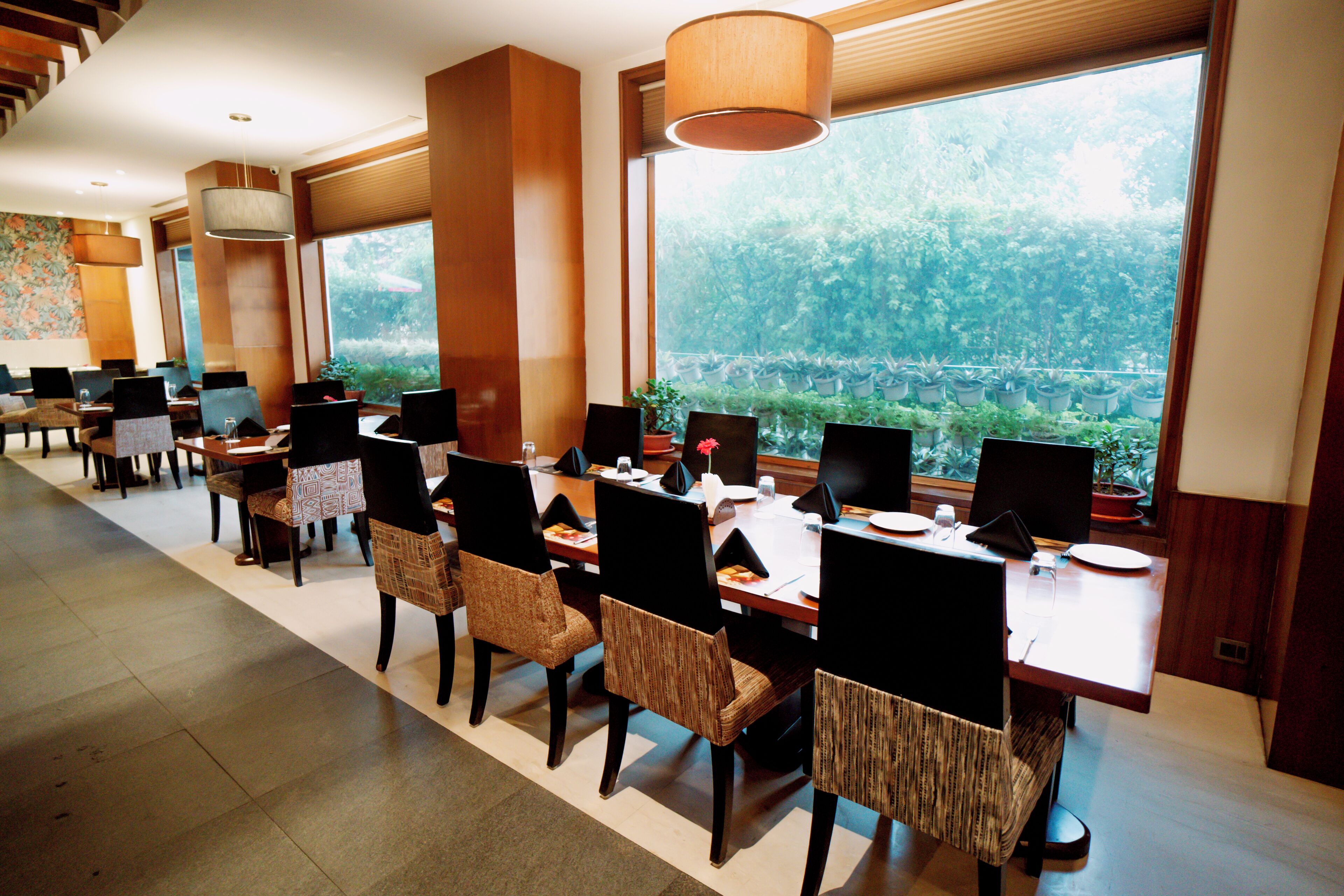A hotel dining area with multiple tables and chairs, large windows with a view of greenery, and pendant lighting.