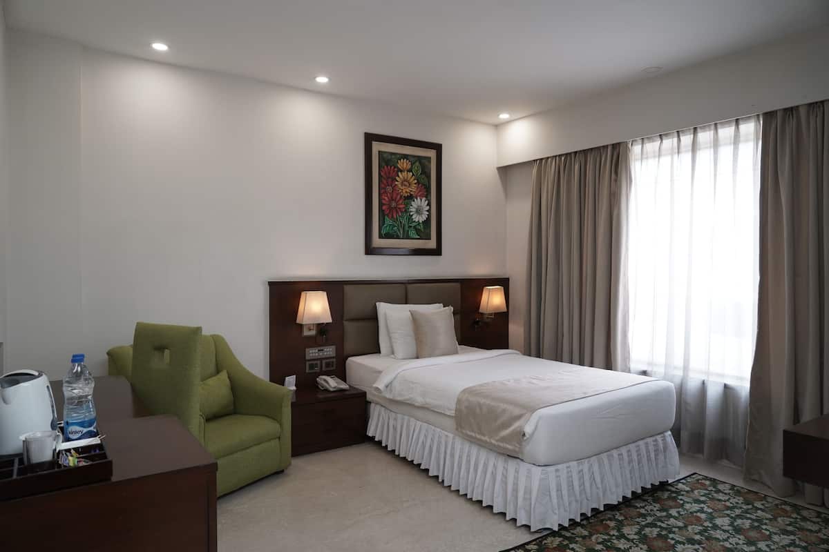Una habitación de hotel con una cama, un sillón verde, un escritorio con hervidor y botella de agua, y un cuadro en la pared.