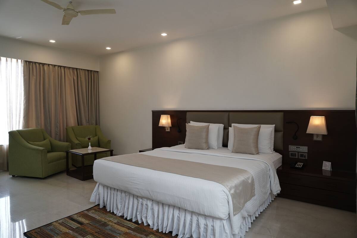 Habitación de hotel con una cama grande, dos lámparas de noche, una mesita de noche, un sillón verde, una pequeña mesa y un ventilador de techo.
