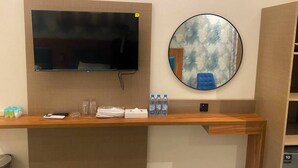 A hotel room with a flat-screen TV mounted on the wall, a round mirror, a wooden desk with toiletries, and a blue chair. - Tamarind Hotel (Dubai)