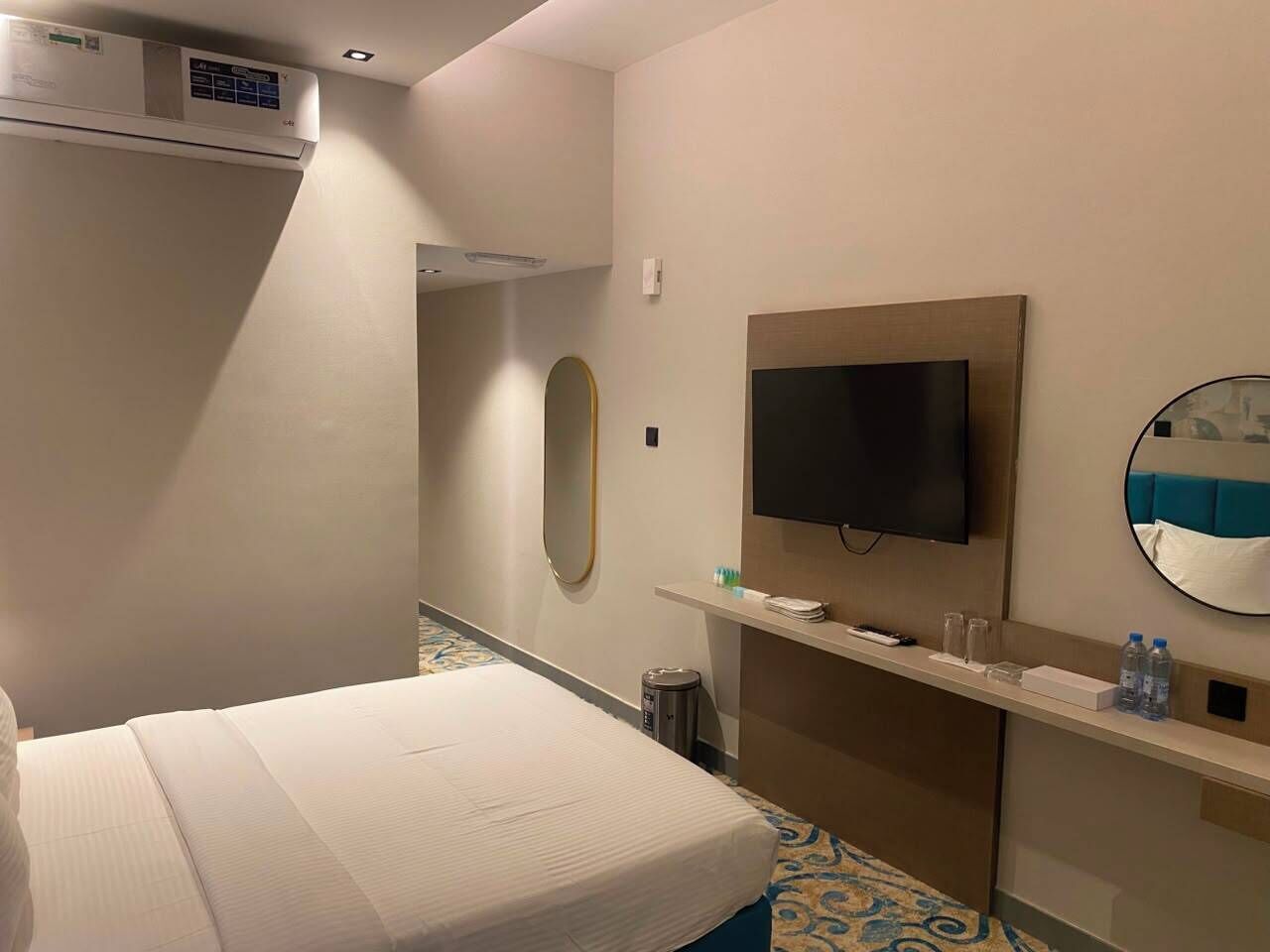 Une chambre d’hôtel équipée d’un lit, d’une télévision, d’un miroir, d’une petite étagère avec des articles de toilette et d’un appareil de climatisation.