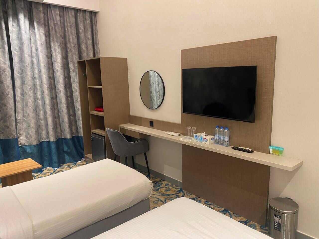 A hotel room with a bed, a desk, a chair, a television, a mirror, and a trash can.