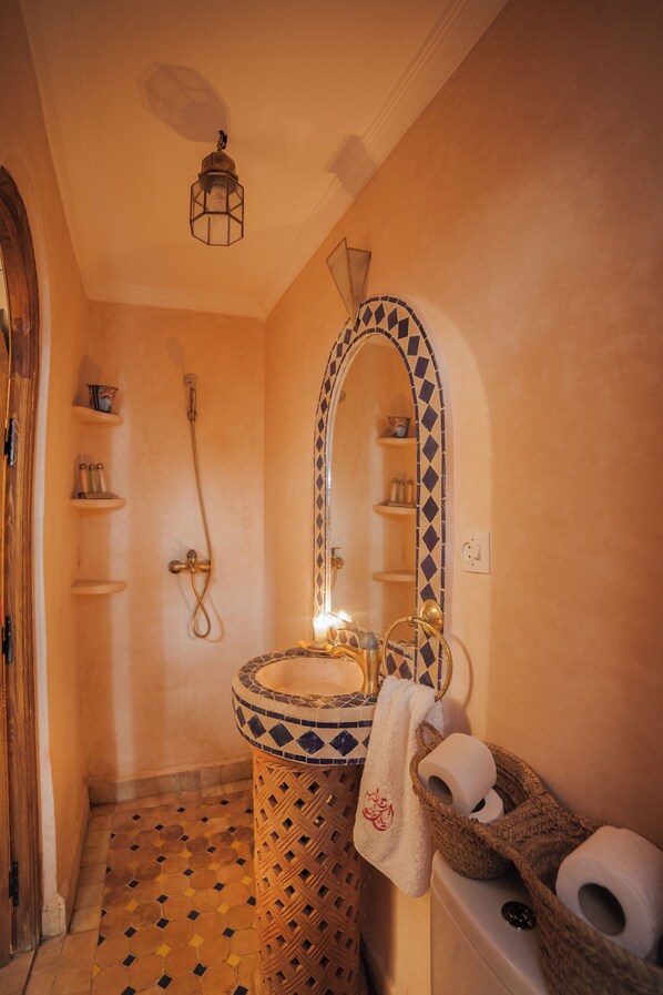 A bathroom with a tiled floor, a sink with a mirror, a towel rack, and a shower area. - Riad Ikalimo 1 (Marrakech)