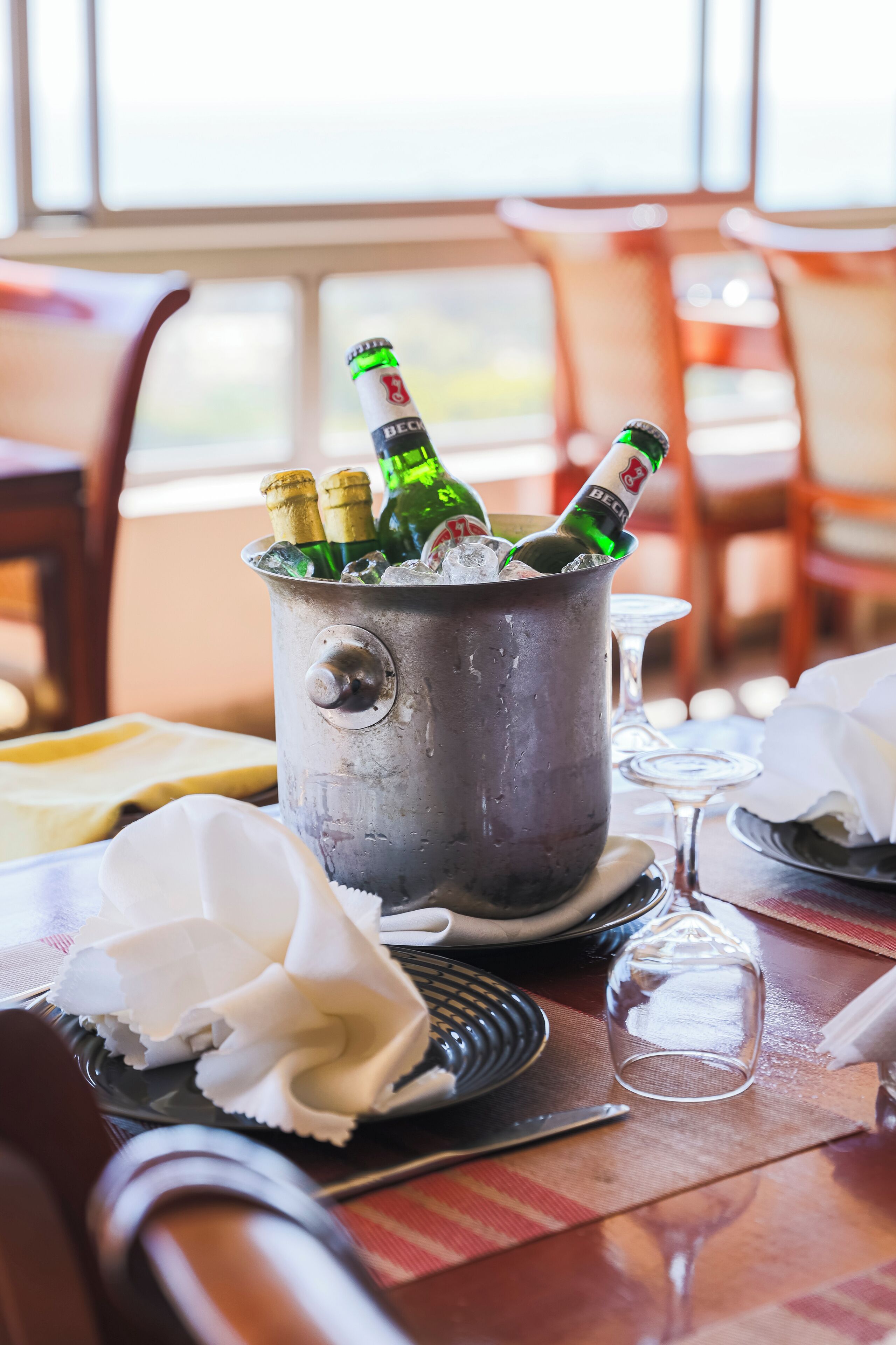 Un seau en métal rempli de bouteilles de champagne et de glace, posé sur une table avec une assiette de nourriture, un verre à vin et une chaise.