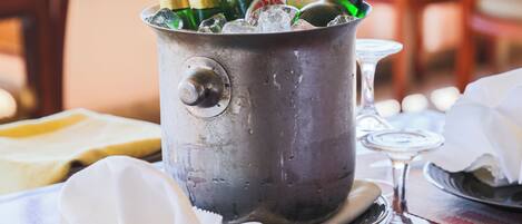 Un seau en métal rempli de bouteilles de champagne et de glace, posé sur une table avec une assiette de nourriture, un verre à vin et une chaise.