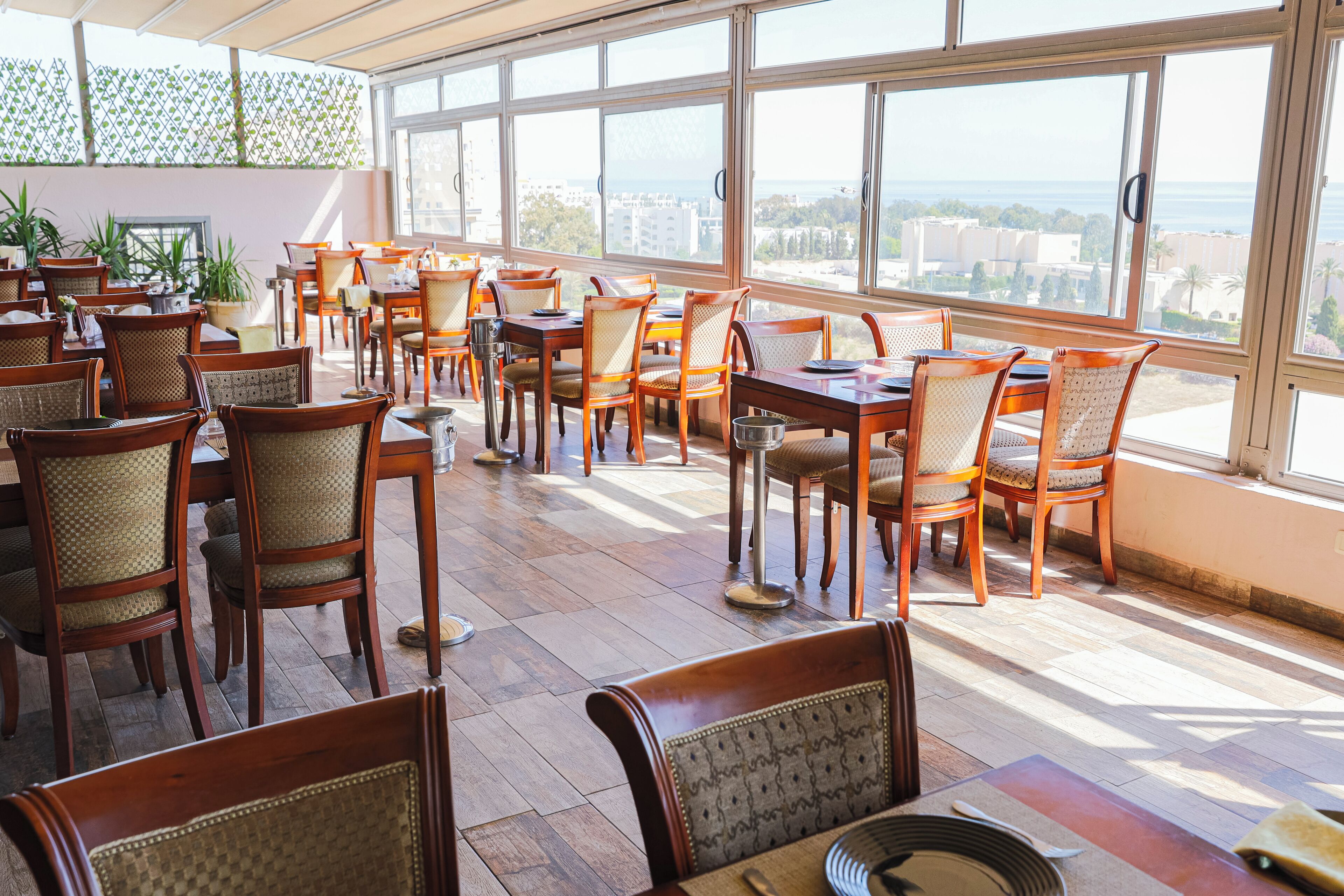 A spacious outdoor dining area with wooden tables and chairs, large windows offering a view of the sea and buildings.