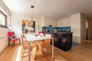 Une cuisine moderne avec des meubles blancs, un îlot central noir et un coin repas comprenant des chaises en bois et une table blanche.
