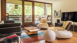 A modern living room with a sofa, ottoman, and a coffee table. There is a TV mounted on the wall and a window with a view of greenery.