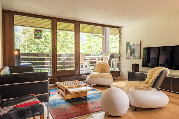 A modern living room with a sofa, ottoman, and a coffee table. There is a TV mounted on the wall and a window with a view of greenery.
