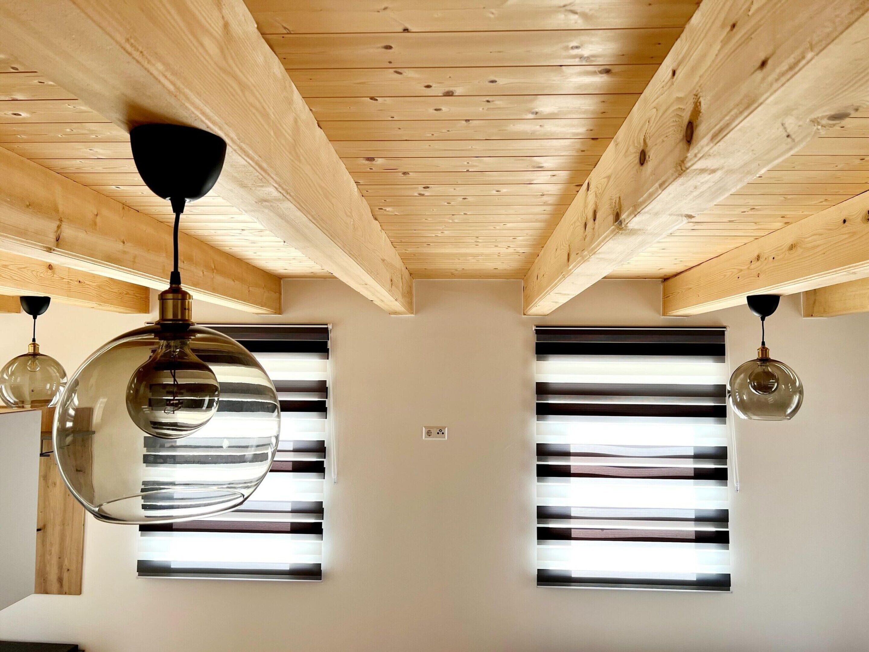 Una habitación con vigas de madera en el techo, dos ventanas amplias con persianas y tres lámparas colgantes.