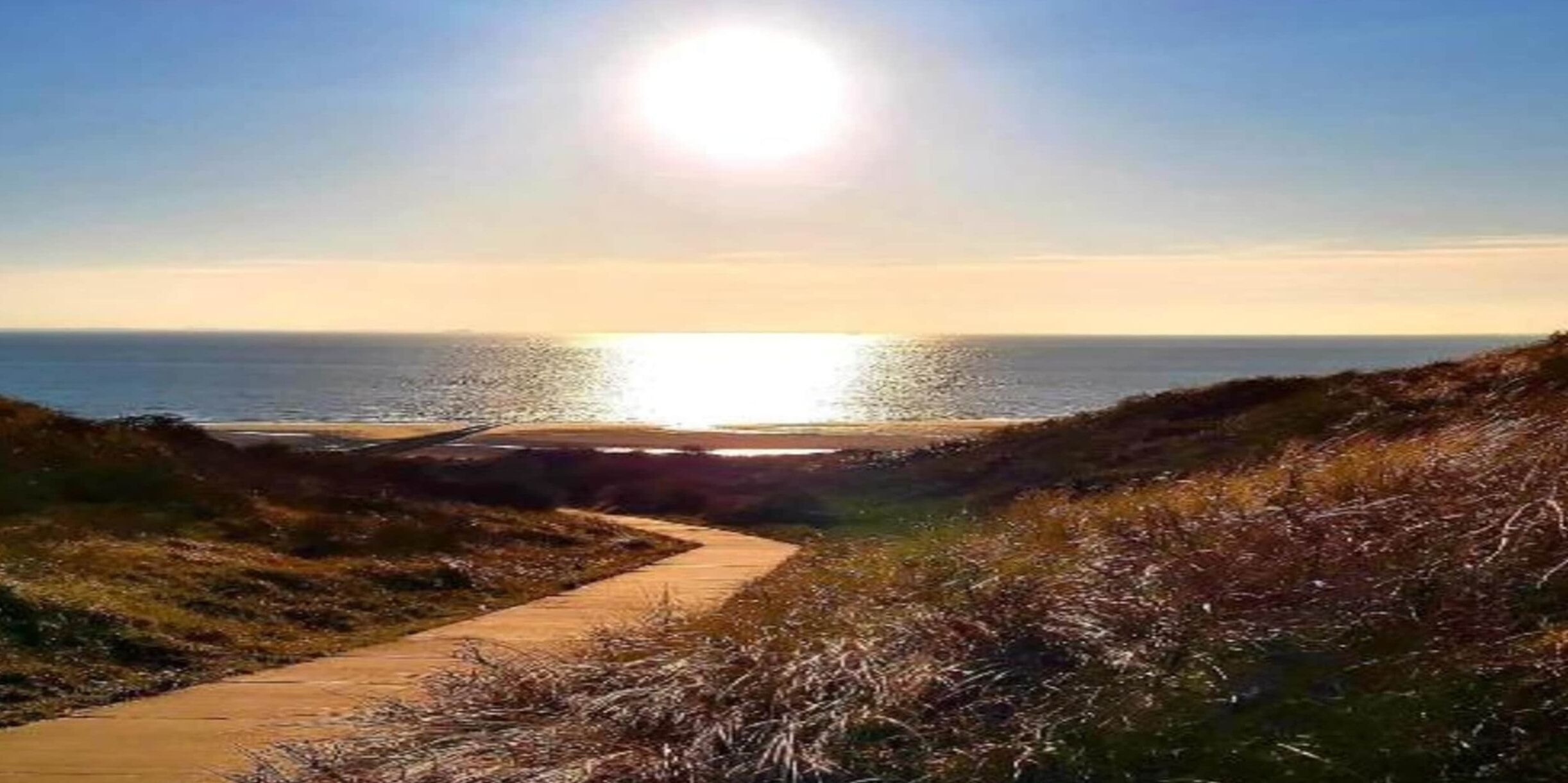 Un sentier côtier menant à la mer, avec un ciel dégagé et un soleil radieux.