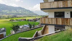 Un edificio moderno a due piani con una terrazza in legno e vista su una valle verde e montagne.
