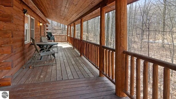 A wooden deck with a railing, a table, and chairs, overlooking a forested area. - Private Wooded Log Cabin Near Shanty Creek Resorts -Four Season Adventure Awaits (Bellaire)
