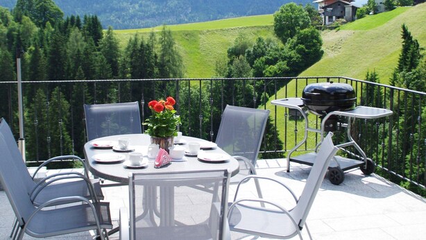 Un balcon avec une table dressée pour les repas, un barbecue et une vue sur une vallée verdoyante et des montagnes.