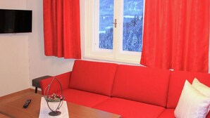 Un canapé rouge, une table en bois avec une télécommande, et une fenêtre avec des rideaux rouges.