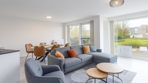 A modern living room with a grey sectional sofa, a round coffee table, and a dining area with chairs and a table. - Corner Apartment in the Center With 3 Bedrooms (Middelkerke)