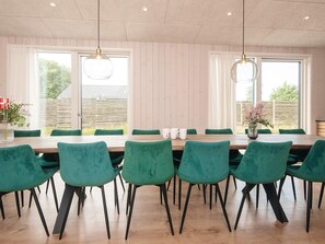 A dining area with a long wooden table and green chairs, large windows with curtains, and hanging pendant lights. - 14 Person Holiday Home in Glesborg (Glesborg)