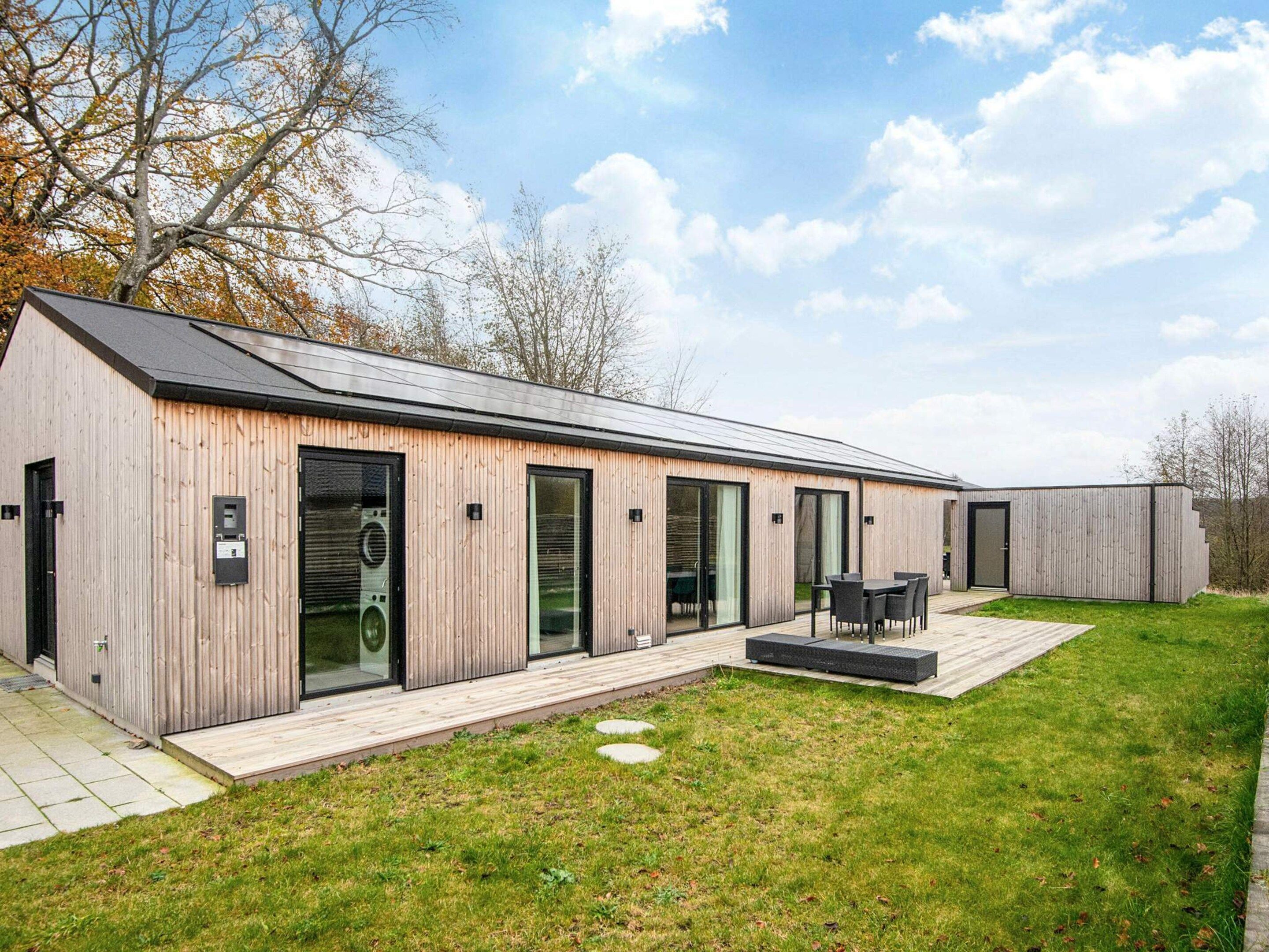 Un bâtiment moderne, de plain-pied, avec une façade en bois, de grandes portes-fenêtres coulissantes et un patio aménagé avec du mobilier d’extérieur.