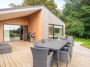 A modern outdoor deck with a dining table and chairs, a grill, and sliding glass doors leading to an interior space. - 14 Person Holiday Home in Glesborg (Glesborg)