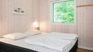 A bedroom with a bed, a bedside table, a wall-mounted shelf with plants, and a window with a view of greenery.
