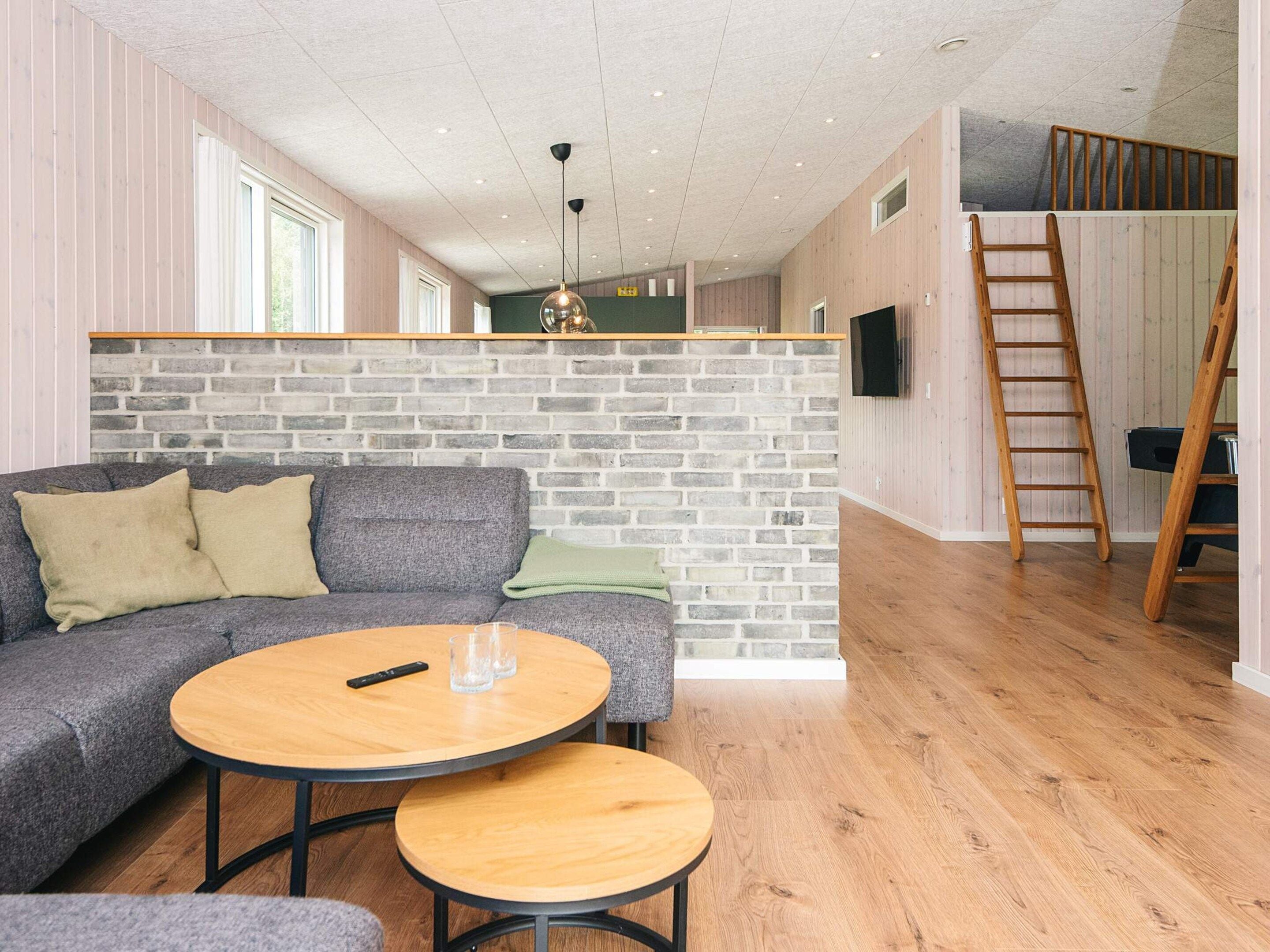 A modern living room with a grey sectional sofa, two round wooden tables, a brick accent wall, and a wooden floor.