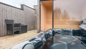 A modern outdoor hot tub area with wooden decking, a glass-enclosed hot tub, and a seating area with wicker chairs. - 14 Person Holiday Home in Glesborg (Glesborg)