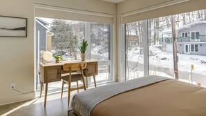 Une chambre avec un grand lit, un bureau avec une chaise, une plante en pot et une vue sur un paysage enneigé par de grandes fenêtres.