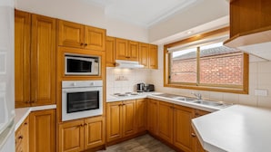A compact kitchen with wooden cabinets, a microwave, an oven, a coffee maker, and a window with a view of a brick building. - Simpson Getaway 1 - Point Lonsdale (Point Lonsdale)