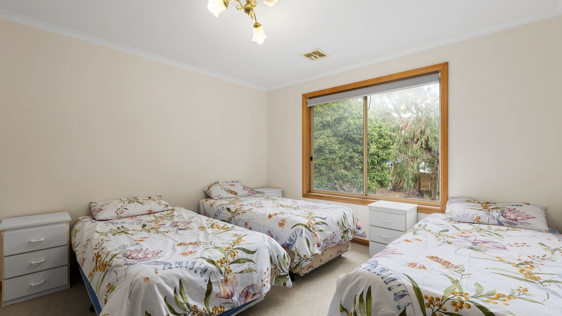 A bedroom with two single beds, a window with a view of trees, a ceiling light, and a small bedside table with drawers.