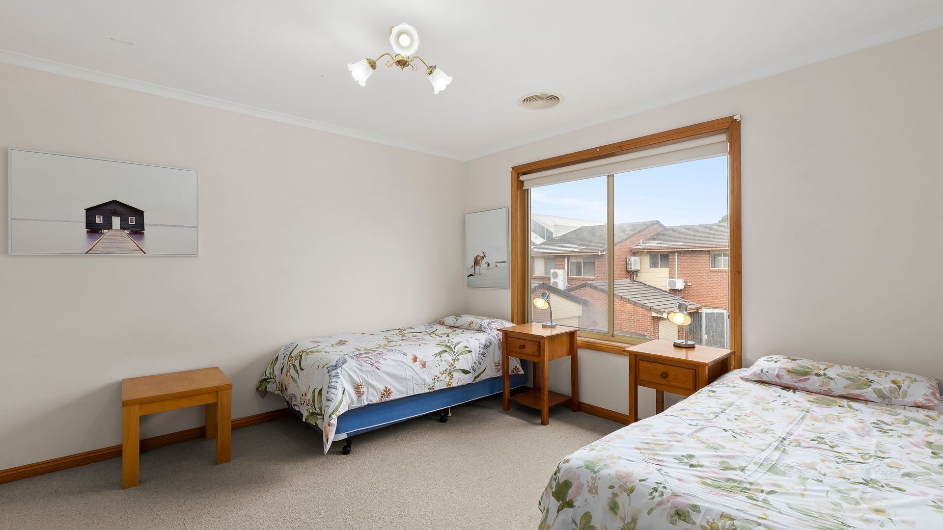 Un dormitorio con dos camas individuales, un escritorio, una silla y una mesita. Se ve una ventana con vistas a casas.