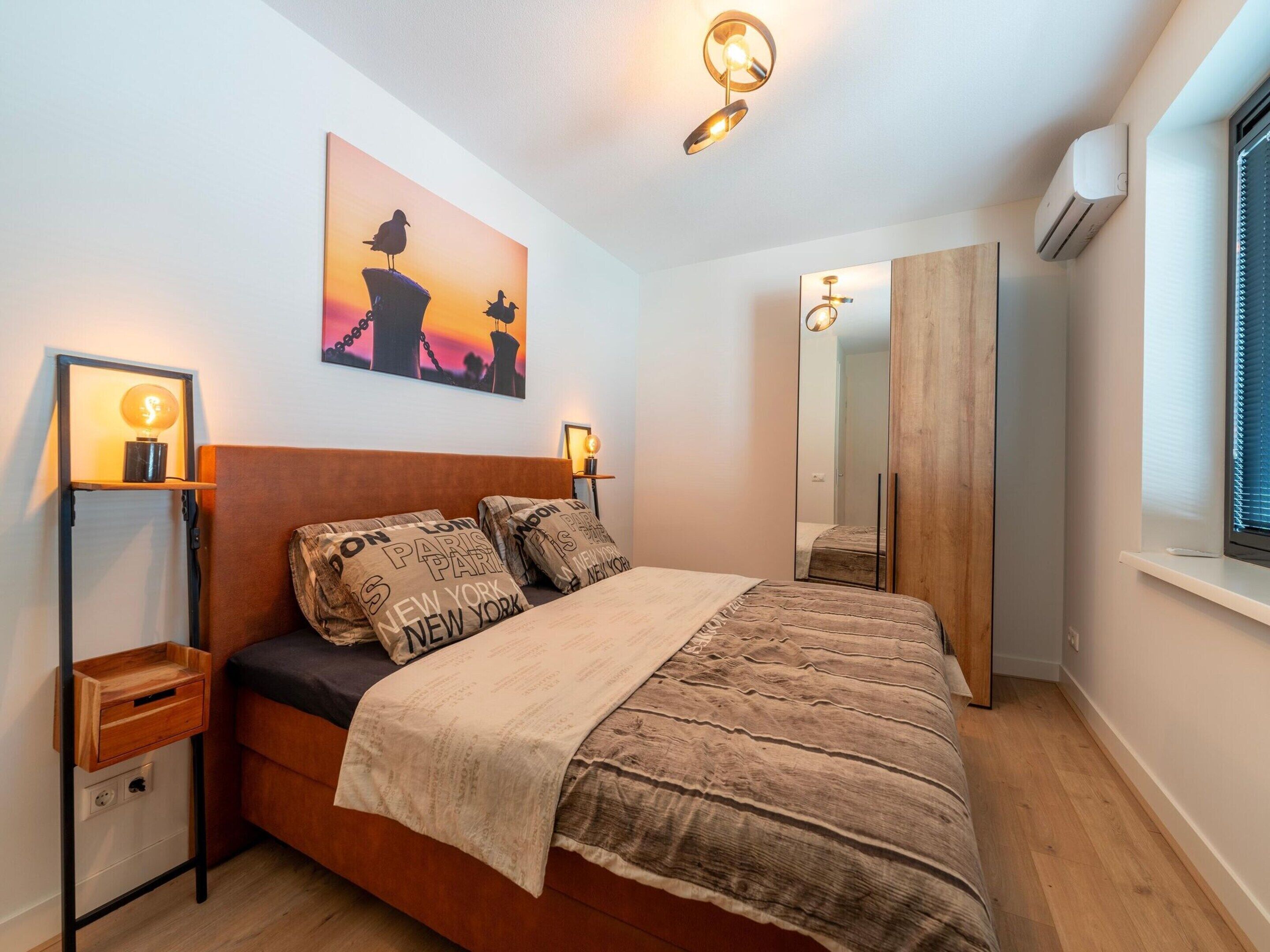 Een slaapkamer met een houten bed, twee nachtkastjes met lampen, een wandairconditioner, een raam met zonwering en een schilderij van vogels op een rots.