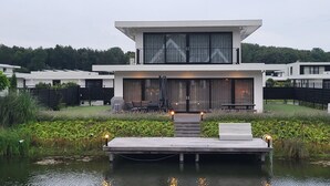 Une maison moderne à deux étages, dotée d’une jetée qui s’avance sur un plan d’eau, et entourée de verdure.