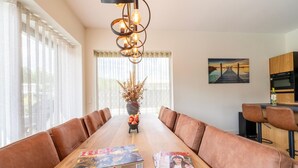 Un espace repas avec une longue table en bois, des bancs recouverts d’un tissu marron, un lustre moderne et un tableau accroché au mur.