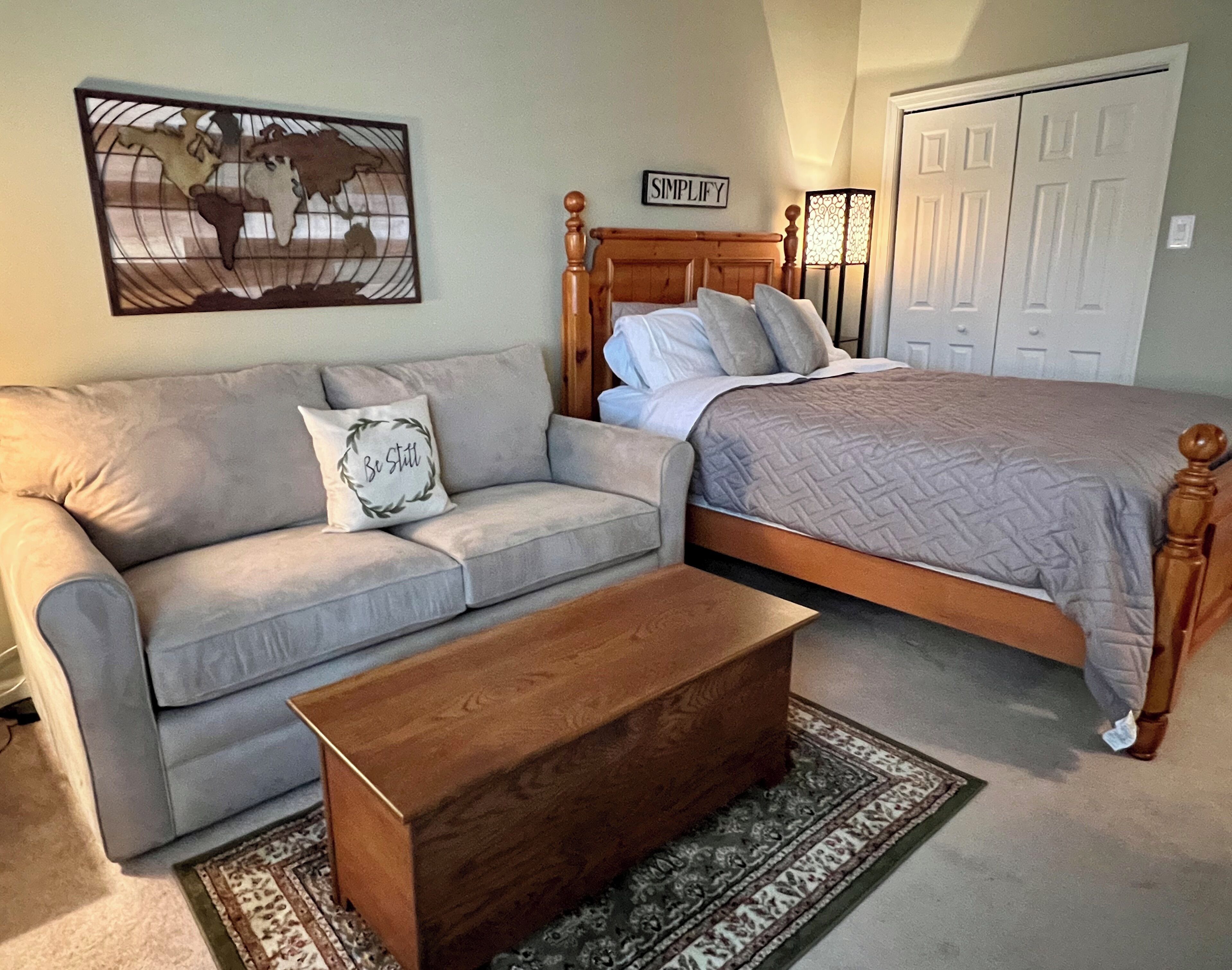 A bedroom with a bed, a wooden headboard, a grey sofa, a wooden coffee table, a patterned rug, a wall art piece, and a door.