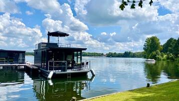 A houseboat docked at a calm lake with a grassy bank, a clear sky, and a distant shoreline with trees.