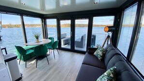 A modern interior with a dining area, a sofa with cushions, and large windows offering a view of the water. - Houseboat Mookerplas With a View (Middelaar)