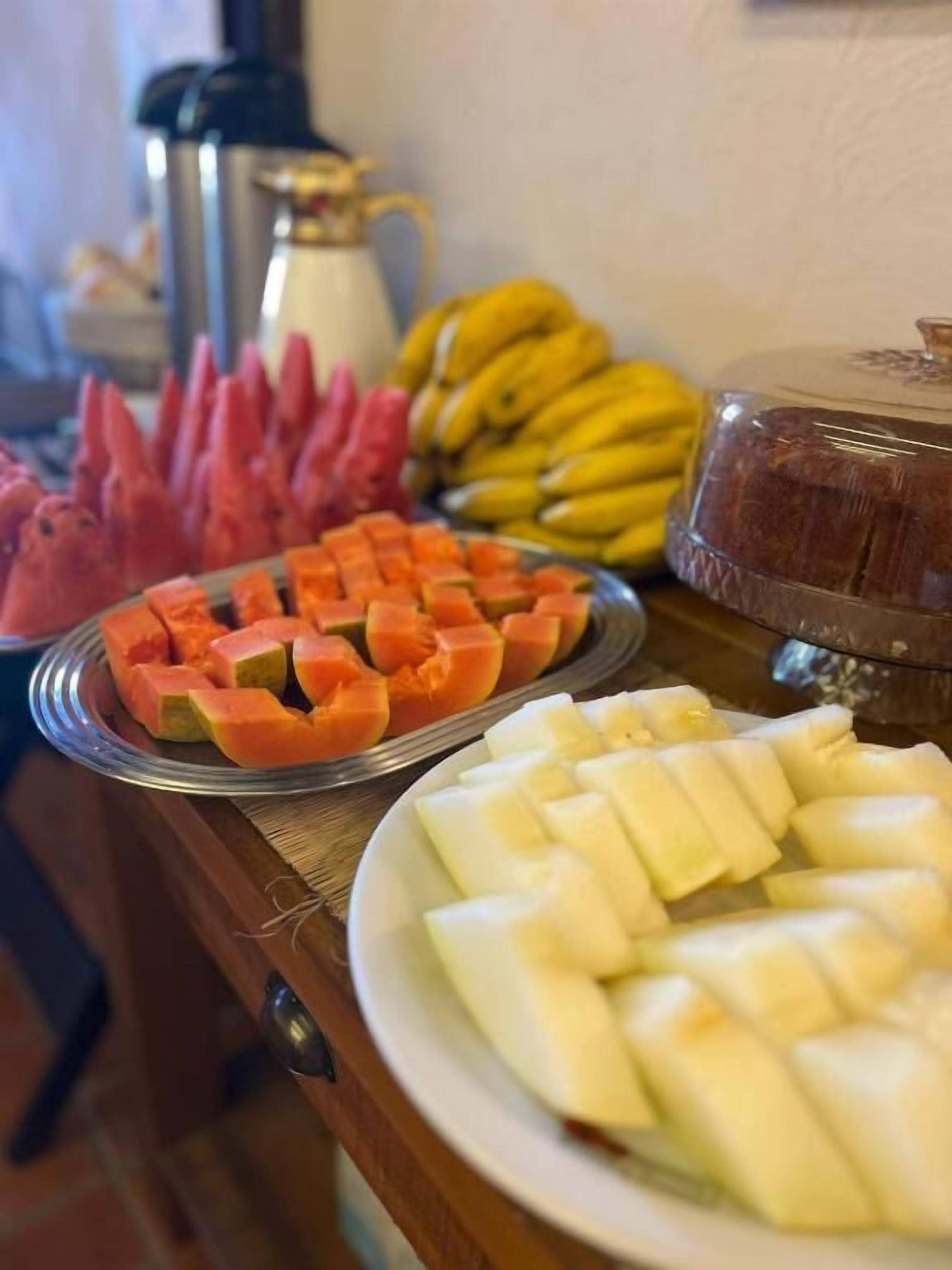 A buffet table with various fruits including watermelon, cantaloupe, bananas, and a loaf of bread.
