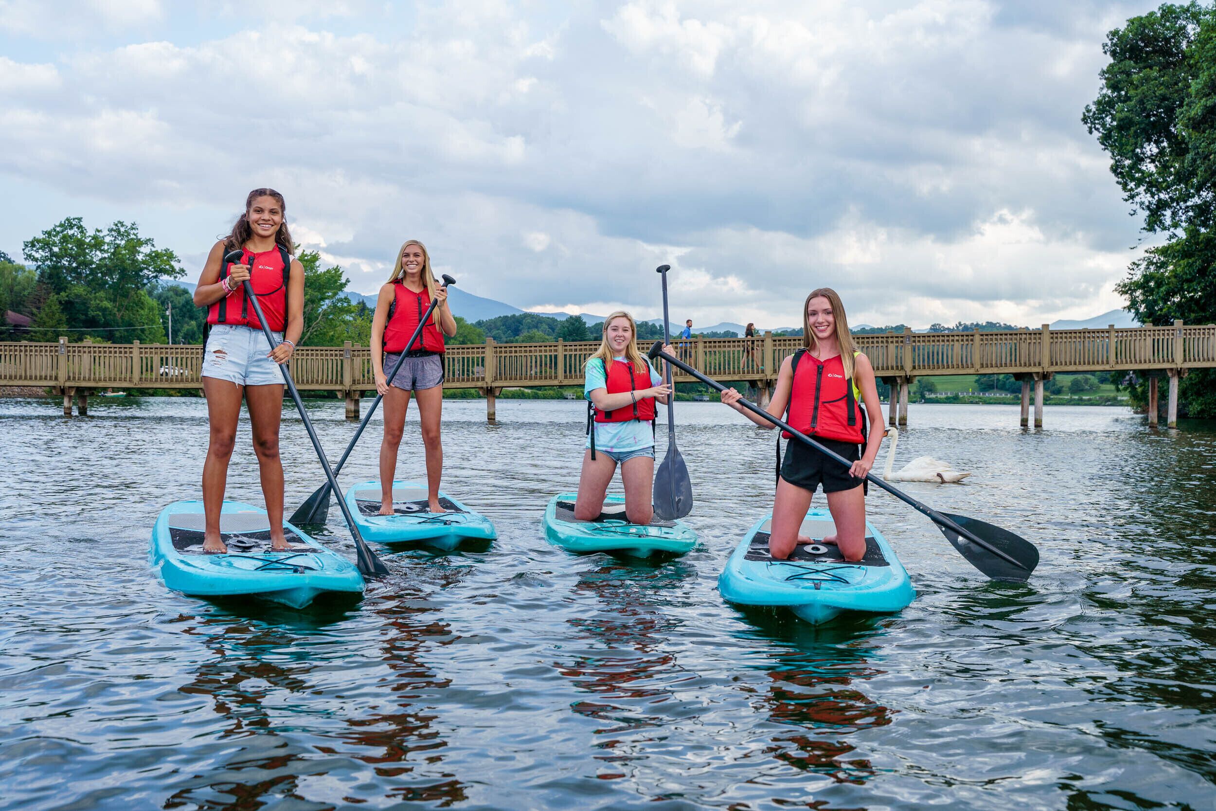 Four people paddleboarding on a calm lake with a wooden bridge in the background.