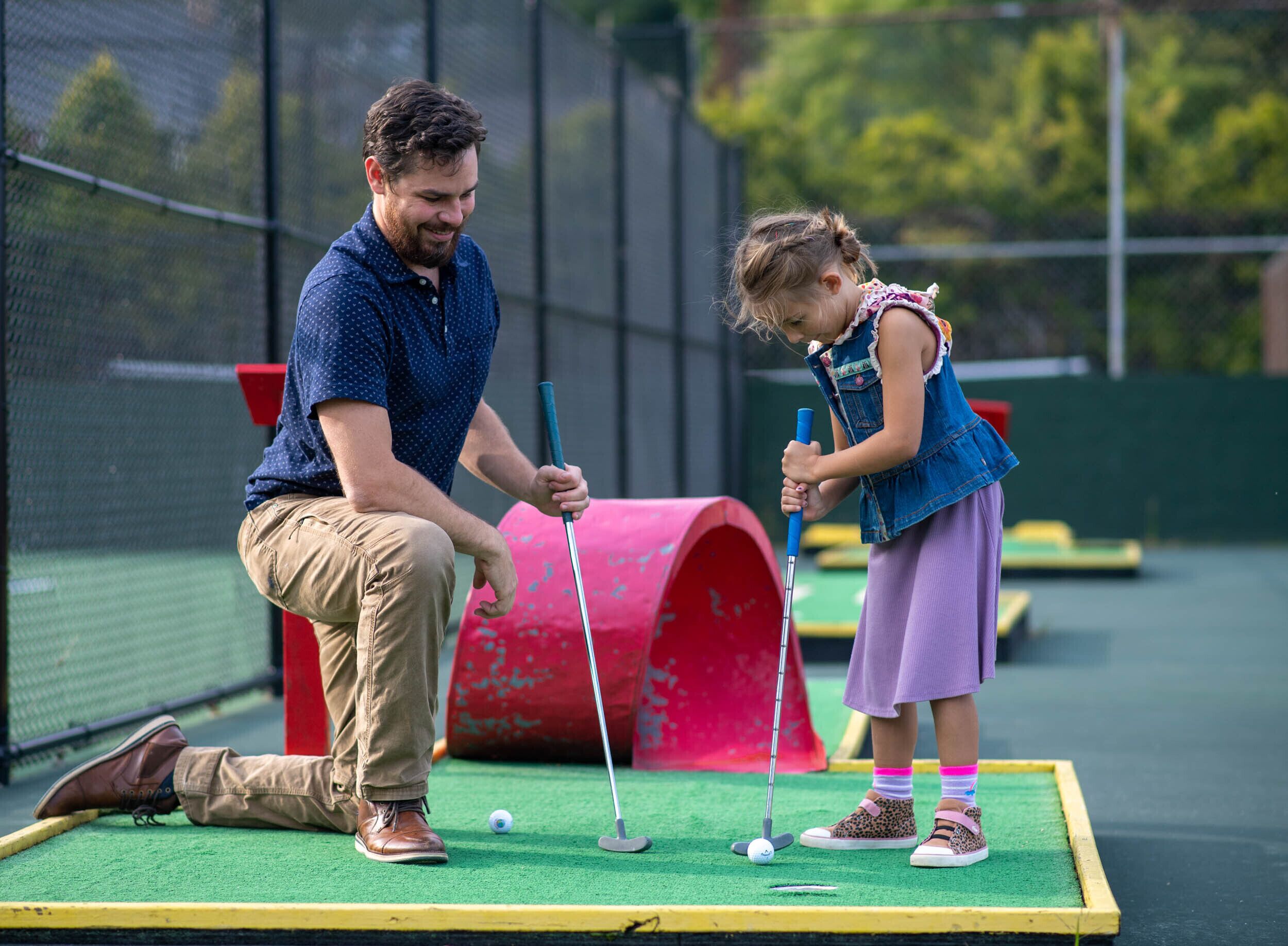 Un hombre y una niña joven jugando minigolf en un campo verde con un obstáculo rojo.