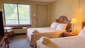 A hotel room with two beds, a desk, a chair, a lamp, and a window with blinds. - Hillside Lodge (Lake Junaluska)