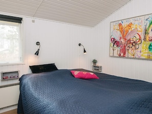 A bedroom with a bed, two wall-mounted lamps, a window, a wall art piece, and a small shelf with a plant.