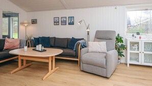 Un salón moderno con un sofá gris, un sillón gris y una mesa de centro de madera. En la pared hay cuadros enmarcados, una lámpara de pie y un aparador con puertas de cristal.