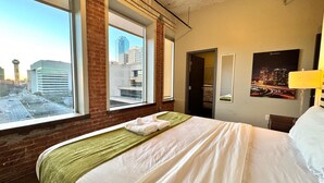 Ein Hotelzimmer mit einem großen Bett, Blick auf die Stadt durch ein Fenster, einem Spiegel an der Wand und einer Lampe.