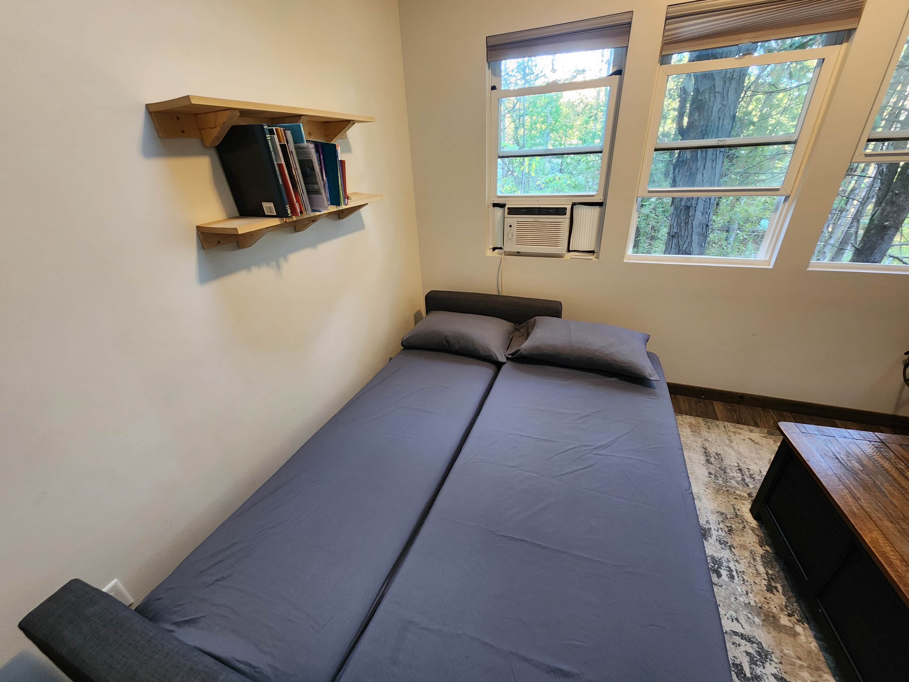 A single bed with a grey mattress and pillows, a wooden shelf with books, and a window with a view of trees.
