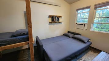 Una stanza con un letto a castello, un letto singolo, una mensola in legno con libri, una finestra vista alberi e un tavolino.