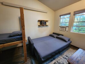 A room with a bunk bed, a single bed, a wooden shelf with books, a window with a view of trees, and a small table. - Tiny Treehouse Cabin | Bluff Woodlands #3 (Elba)